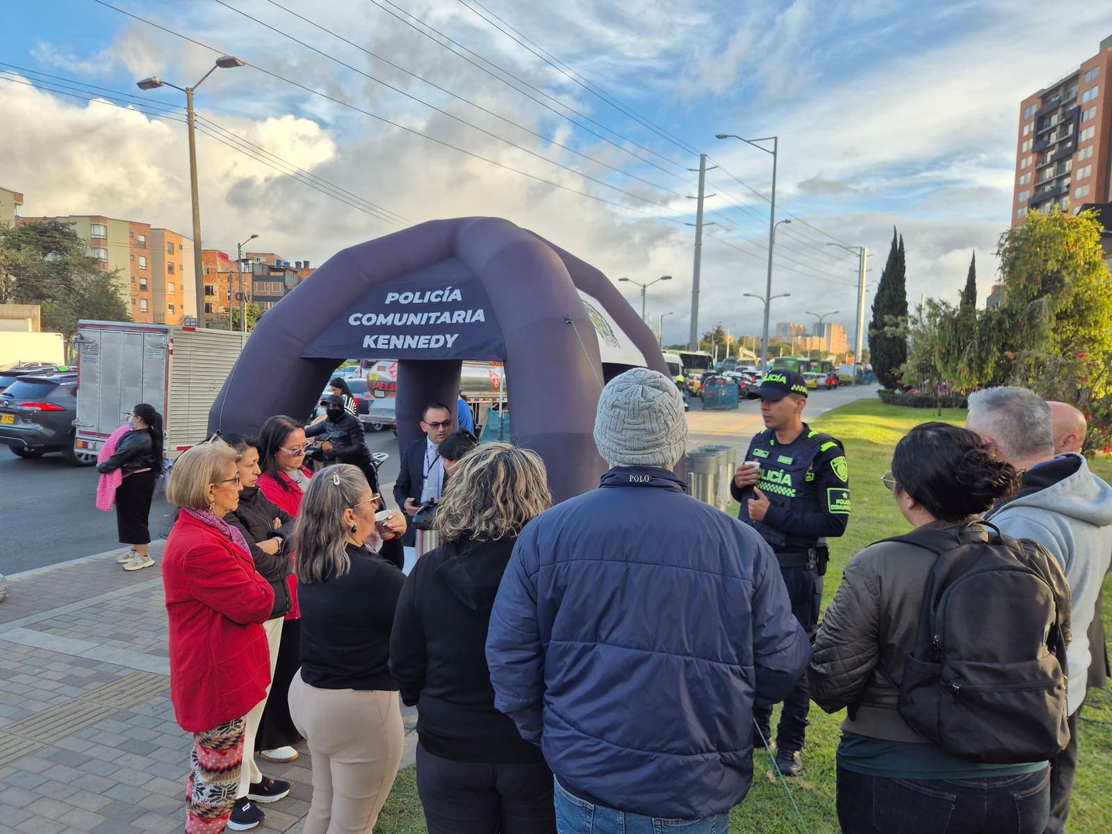 Vecinos escuchando a los patrulleros junto al arco inflable de la Policía Comunitaria de Kennedy, con el tráfico de la Avenida Carrera 68 al fondo