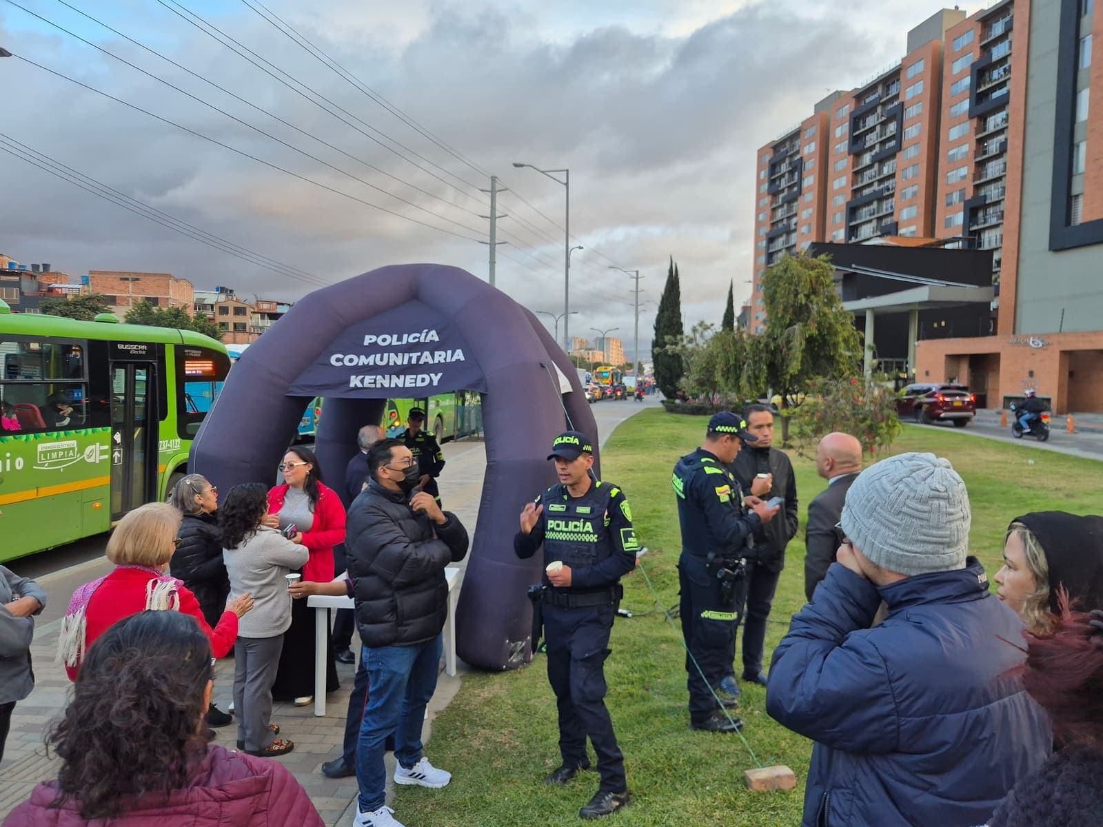 Patrulleros de la Policía Comunitaria y residentes conversando en la zona verde, con un bus del sistema de transporte al fondo sobre la Avenida Carrera 68